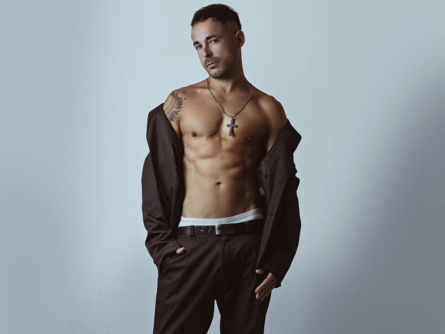 AdamsSanz, shirtless, with a toned, muscular, brown-skinned body, black pants, hands in pockets, cross necklace, tattoo on right shoulder, short brown hair, serious expression, standing.