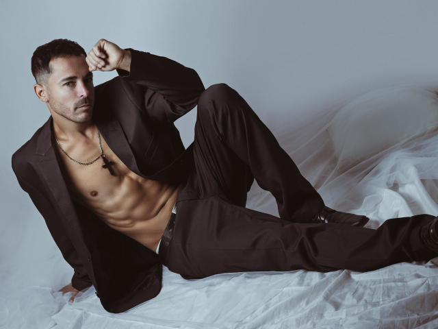 AdamsSanz with a lean, muscular build, wearing an open black suit, exposing his abs, lying on white sheets, looking at the camera with a slight smirk.