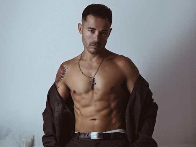 AdamsSanz with a toned, shirtless, muscular upper body, black jacket off, cross necklace, tattoo on right shoulder, and a serious expression. His ass is partially visible.