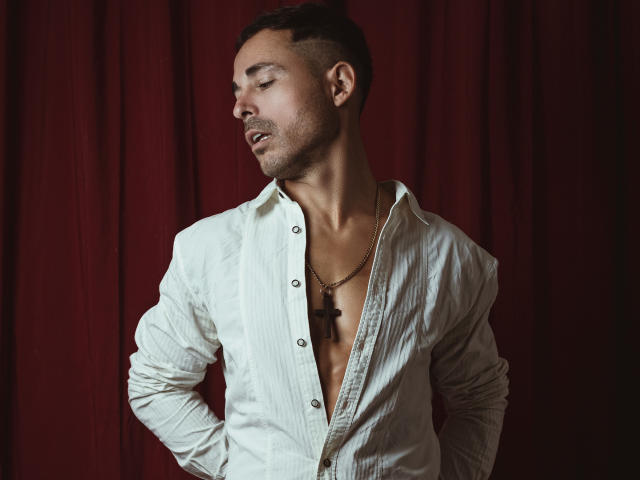 AdamsSanz with a serious face, eyes closed, wearing an unbuttoned white shirt revealing a necklace, hands in pockets, short dark hair, red background.