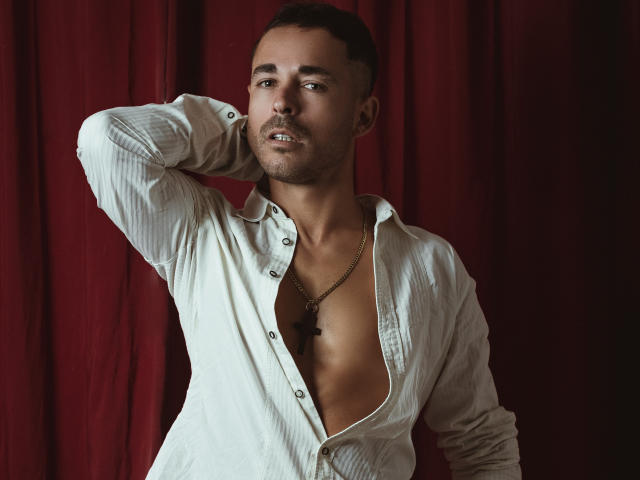 AdamsSanz with short brown hair, wearing an unbuttoned white shirt revealing a bare chest and a cross necklace, has one arm behind his head, looking slightly to the side.