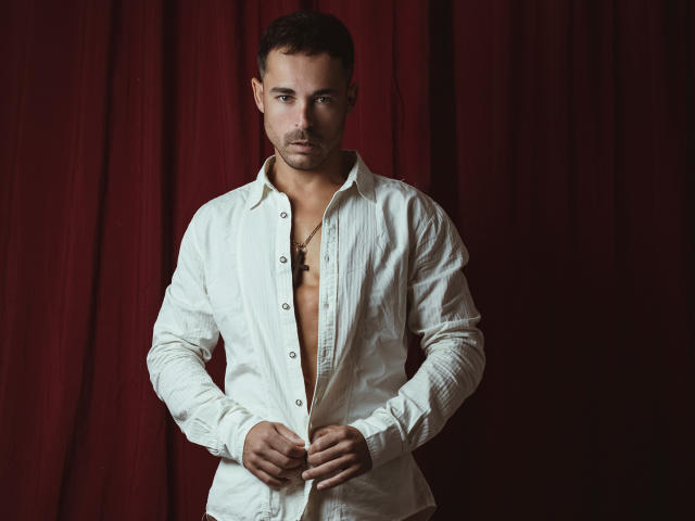 AdamsSanz with short brown hair, slight beard, and intense look, wearing an unbuttoned white shirt, exposing a small tattoo on his chest, against a red curtain.
