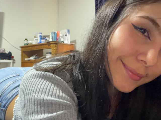 Close-up of SarahPrincess with a slight smile, dark straight hair, and subtle winged eyeliner. She's wearing a gray ribbed shirt and denim shorts. Her face is in the foreground, slightly angled. Background shows a wooden shelf with various items. She's lying down, her head to the right, upper body to the left. Skin is light, lips are pink. The image has a casual, everyday vibe. SarahPrincess's expression is relaxed and confident.