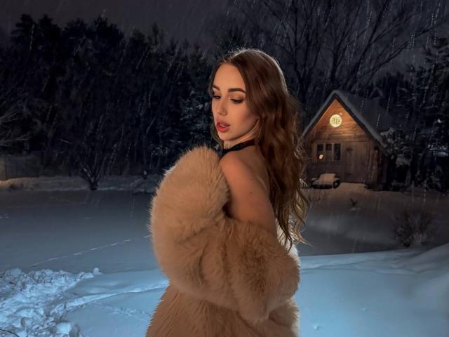 IsabelleDelicious with long brown hair, wearing a fluffy beige coat, looking over her shoulder, snow in the background, standing in front of a wooden house with a lit window.