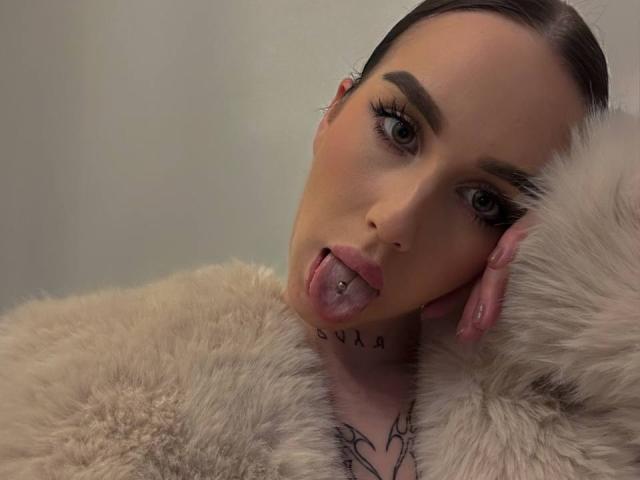 Close-up of IsabelleDelicious with light skin and dark eyebrows, tongue out with a small tongue piercing. She has blue eyes, long eyelashes, and a subtle blush. IsabelleDelicious wears a beige fur coat with a fluffy texture, covering her shoulders. Her right hand is resting on the coat, with pink, glossy nails. A black tattoo with heart shapes is visible on her chest. Her dark brown hair is pulled back. The image has a soft, muted light. IsabelleDelicious's expression is playful and slightly provocative. The overall composition is simple, focusing on her facial features and the texture of the fur coat.