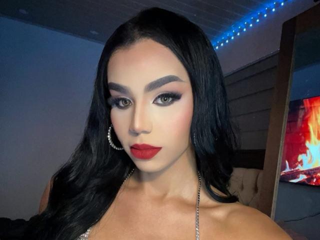 Portrait of OriannaSimons. She has long black hair, wearing bold makeup with dark, defined eyebrows, and bright red lipstick. Her eyes are lined with thick, black eyeliner, and she wears large silver hoop earrings. She has a light skin tone, and her expression is neutral. She's wearing a silver chain necklace. The background has blue LED strip lights and a TV displaying a fiery scene. Her makeup and appearance are sharply defined, emphasizing her eyes and lips. The image is sharply focused, highlighting her facial features.