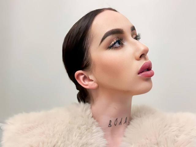 IsabelleDelicious with puffy pink lips, dark slicked-back hair, and a fluffy white coat, has "BOOM" tattooed on her neck, looking up with half-closed eyes and long lashes.
