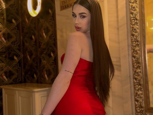 IsabelleDelicious with long brown hair, red strapless dress, tattoo on upper arm, looking over shoulder, standing in dimly lit room, yellow light, ornate mirror.