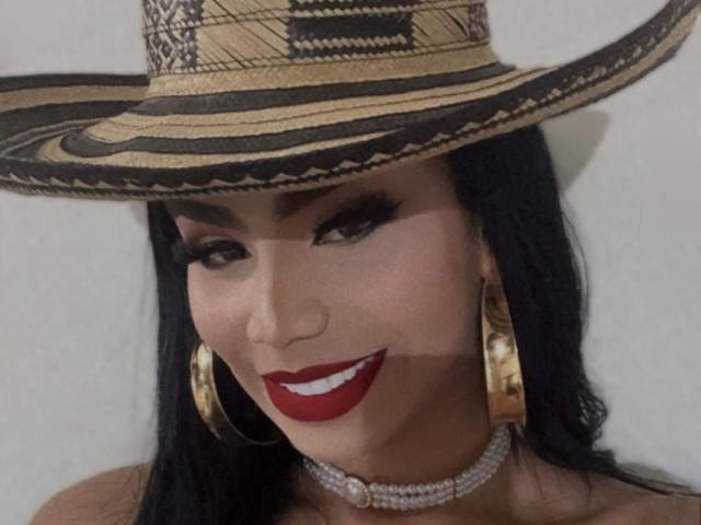 OriannaSimons has long black hair, wearing a large woven straw hat with black and gold patterns. She has dark eyebrows, thick eyeliner, and bright red lipstick. Her gold hoop earrings and sparkly choker contrast with her smooth, tan skin. Her smile is confident and she looks directly at the viewer. The composition is tight, focusing on her face and hat. The texture of the hat is detailed, with visible weaving patterns.