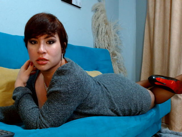 SaraKleine is lying on a blue couch, wearing a grey knit dress, black stockings, and red high heels, with her ass up and looking at the camera.