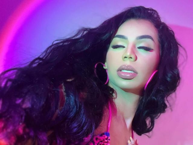 OriannaSimons with long black wavy hair, pink hoop earrings, and glossy lips, eyes closed, wearing a colorful top, bathed in pink and purple neon light.