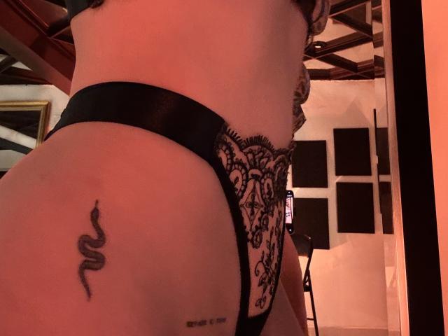 HannaBorbons Hintern in schwarzen Spitzenunterwäsche, wobei ein Schlangentattoo auf ihrer rechten Gesäßwange zu sehen ist, mit einem Spiegel und einem Telefon im Hintergrund. Warmes, rotes Licht..