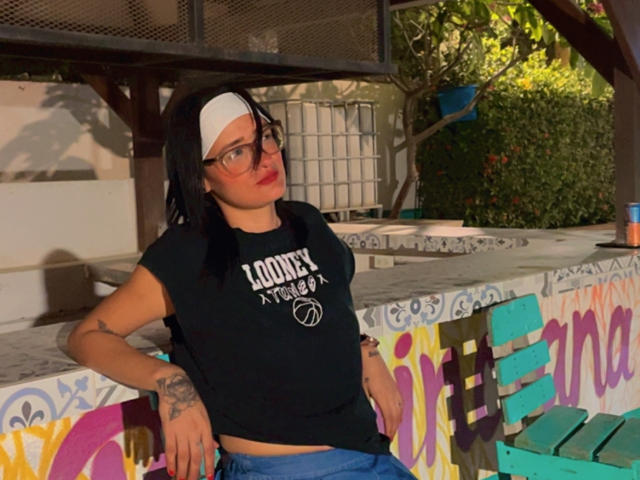 CattaleyaEvans, black t-shirt, white headband, glasses, tattoos, leaning on colorful graffiti wall, blue pants, red lips, green eyes, black hair, casual outdoor setting.
