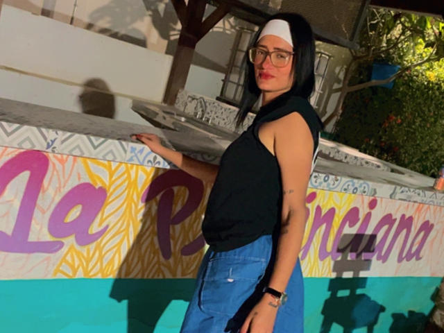 CattaleyaEvans with black bob, glasses, white headscarf, black sleeveless top, blue pants, leaning on wall, smiling, short brown hair, small tits, medium build, standing by pool.