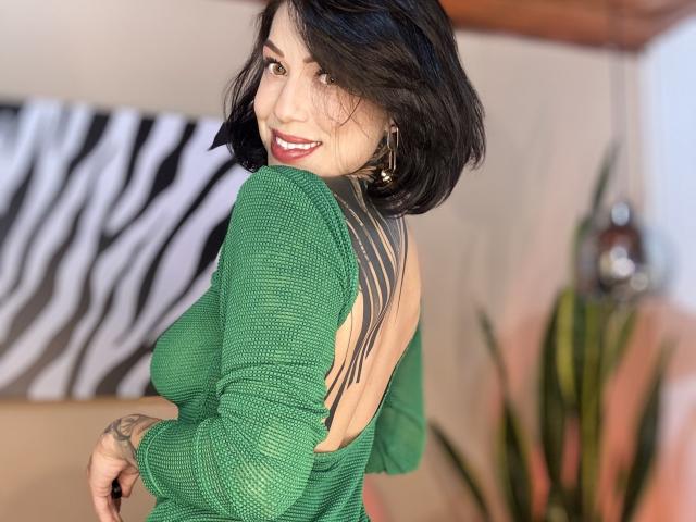 HannaBorbon with short black hair, green knitted top, open back showing her tits, smiling at the camera, tattoo on right shoulder, green plant in background.