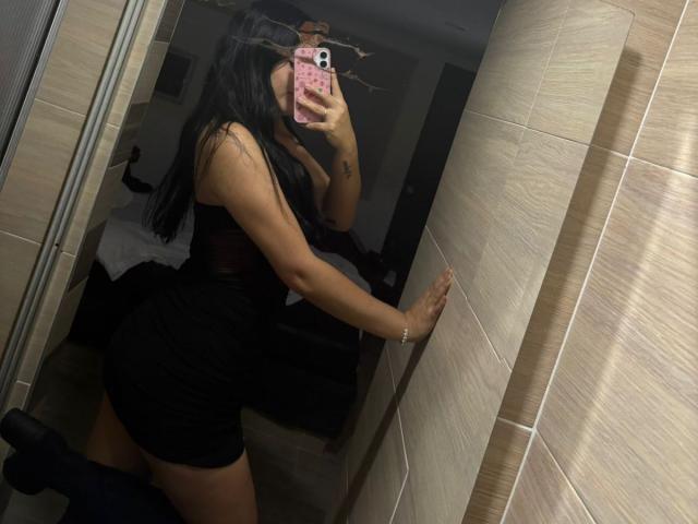 DulceOh69 with long black hair, wearing a black dress, taking a mirror selfie, showing her ass, holding a pink phone, against a tiled wall in a dim room.