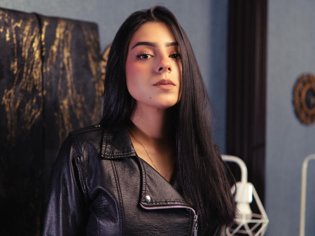 ZoeBellini has long black hair, wearing a black leather jacket, with a small mole on her right cheek, looking slightly to the side with a serious expression.