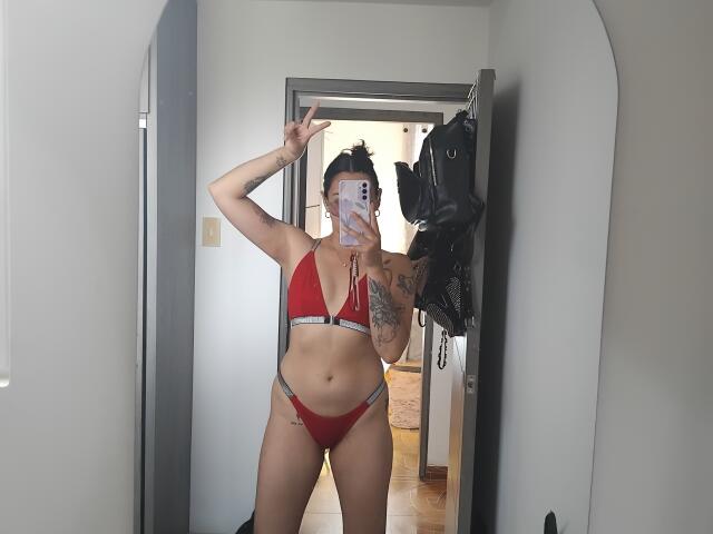 Mllevv stands in a mirror, wearing a red bikini with black trim. She has tattoos on her right arm and left hip, and her dark hair is in a bun. She holds a phone with a white case in her right hand, making a peace sign with her left. A black leather jacket hangs on a hook in the doorway behind her. Her skin is light, and she has a slim, toned physique. The mirror reflects a wooden floor and a partially visible room.