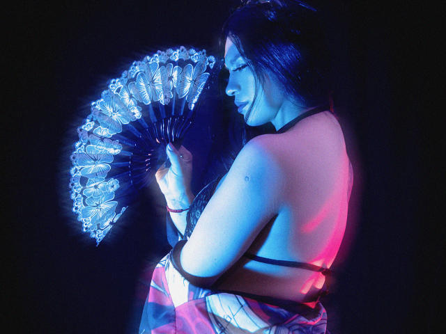 GreizSubKimura con illuminazione al neon blu e rosa, che tiene una ventola a forma di farfalla luminosa, di spalle, capelli neri, spalle scoperte, tatuaggio sul braccio destro, tessuto rosa e bianco scintillante..