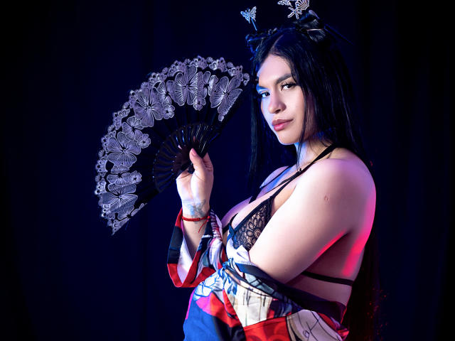 GreizSubKimura with long black hair, wearing a kimono, holding a fan with butterfly patterns, showing her tits, and a tattoo on her left arm, against a dark background.