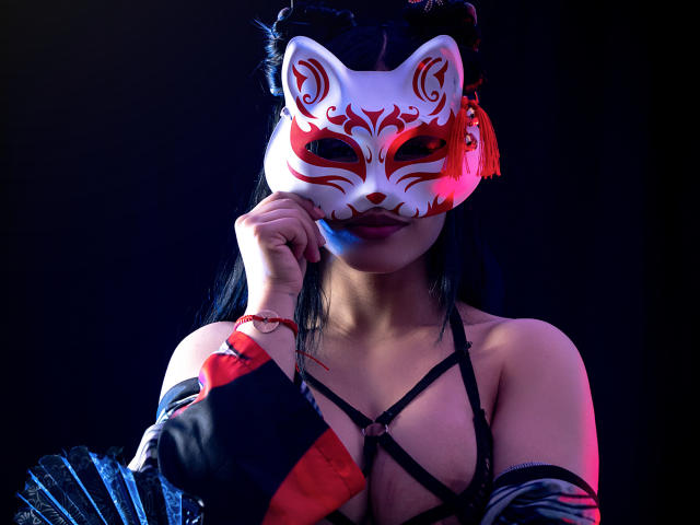 GreizSubKimura wearing a white fox mask with red markings, black hair, holding mask with right hand, black strappy top, red and black sleeves, dark background, purple and blue lighting.