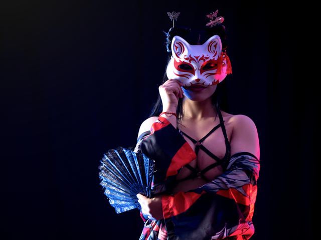 GreizSubKimura wears a red and white fox mask, black strappy top, and holds a blue fan. She has black hair, red and black clothes, and a seductive pose.