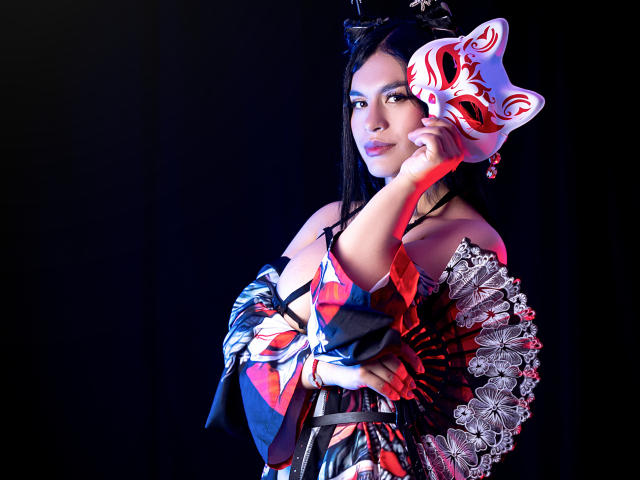 GreizSubKimura with black hair, wearing a colorful kimono, holding a red and white mask, and a lace fan, against a dark background. She's looking at the camera.