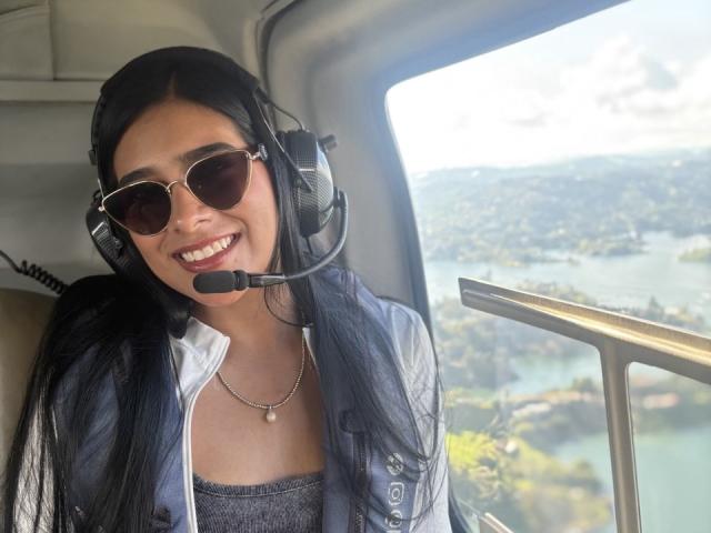 AntonellaPreschot with long black hair, wearing a headset, aviator sunglasses, and a blue jacket, smiling, showing her teeth, and looking out of a window. Her tits are not visible.