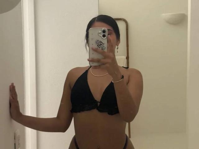 AntonellaPreschot with black bra covering her tits, holding a phone in front of her face, wearing a gold necklace, standing in a bathroom with white walls.