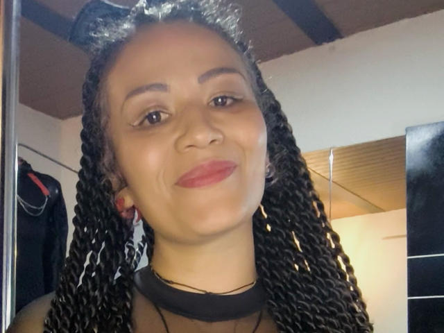 Close-up photo of DivaMarie with medium brown skin and black braided hair. She's wearing a black choker and cat ear headband. She has red lipstick and subtle eye makeup. DivaMarie's eyebrows are dark and well-defined. She's smiling slightly, showing a small dimple. The ceiling has wooden beams and the room has white walls. Her earlobes have red earrings. The lighting is even, highlighting her face. The composition focuses on her upper body. Slightly tilted, she's centered in the frame.