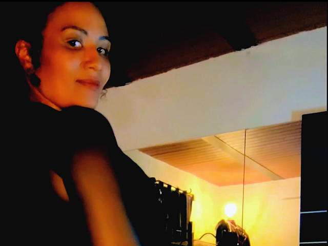 DivaMarie with dark hair, black ears, and a slight smile, wearing a black top, in a dimly lit room with a warm light in the background.