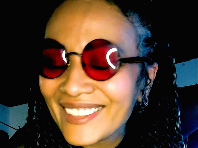 DivaMarie with red, reflective sunglasses, smiling, wearing black braids, her brown skin glowing under a blue light. Her white teeth are visible, and she's looking slightly to the side.