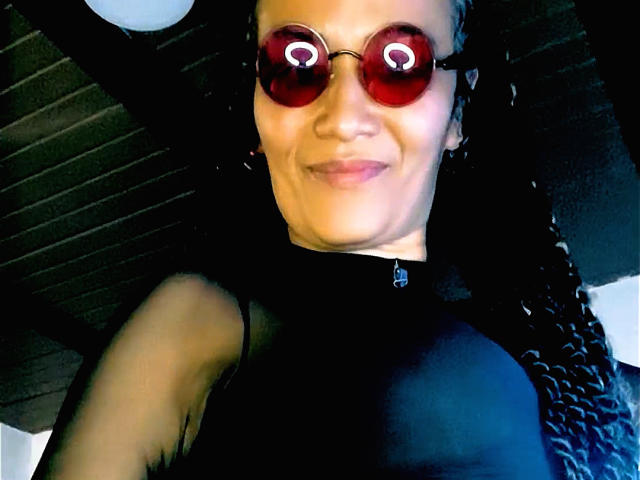 DivaMarie with red reflective sunglasses, smiling, wearing a black top, showing her tits, with braided hair, in a low-angle selfie.
