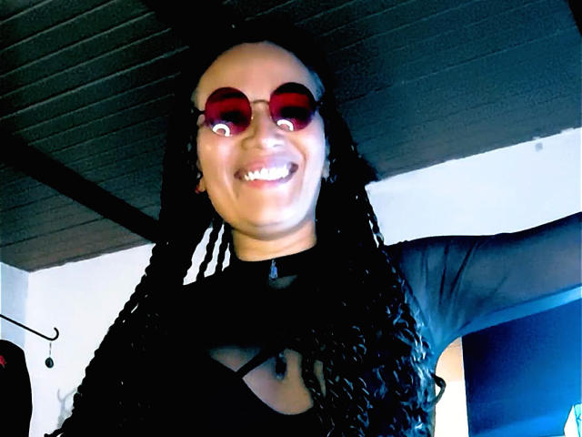 DivaMarie with long black braids, wearing red circular sunglasses, smiling with her eyes winking, in a black sheer top, under a dark wooden ceiling.