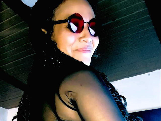 DivaMarie with black braids, wearing heart-shaped red sunglasses, and a black sheer top, showing her bare shoulder and a small tattoo, smiling slightly.