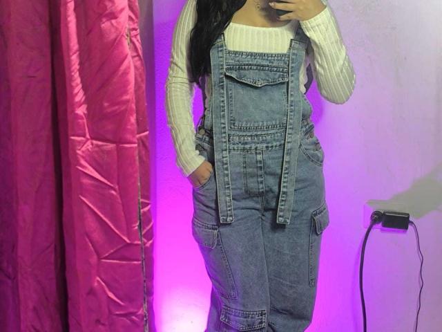 LissyCarter wearing blue denim overalls with white ribbed shirt, hands in pockets, purple and pink background, visible black phone charger.