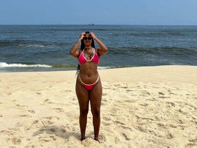 NoirSeduction69 stands on a sandy beach, wearing a pink bikini, holding sunglasses, with dark skin, long black hair, and a necklace, looking at the camera.