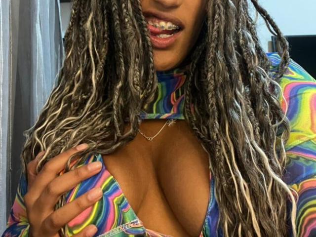 NoirSeduction69 with long blonde and brown braided hair, wearing a colorful, tie-dye top, showing her cleavage, smiling with visible teeth, hand on her chest.