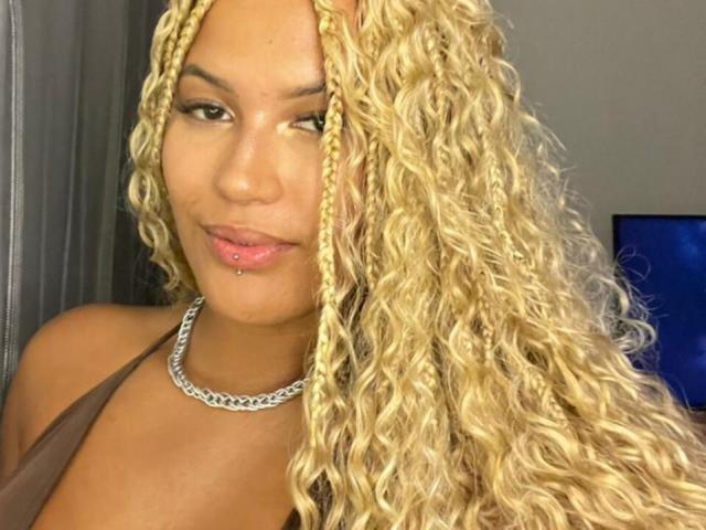NoirSeduction69 has long blonde curly braids, a brown halter top, a silver chain necklace, and a pink lip piercing, smiling at the camera.