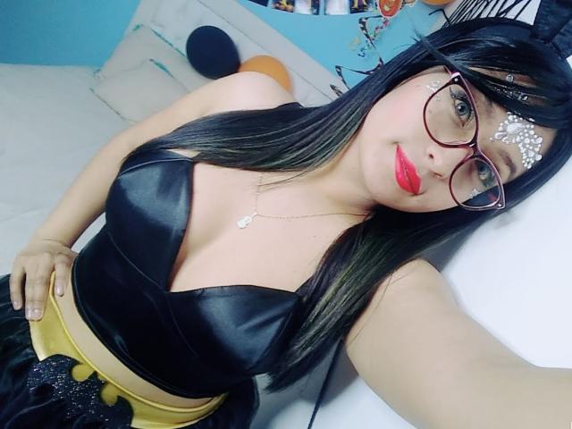 SofiiaThooner with black hair, glasses, white face paint, red lipstick, black leather top, yellow skirt, hand on hip, sitting on white couch, looking at camera.