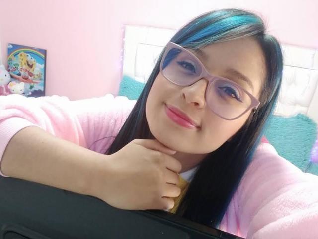 SofiiaThooner with glasses, pink cardigan, and black hair with blue highlights, smiling softly, hands resting on a black surface, pink lipstick, and light pink wall in background.