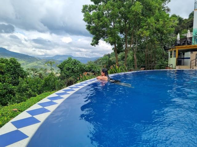 SofiiaThooner's ass is above the water in a blue, curved pool, with a tree-lined, mountainous view in the background, and a house on the right.