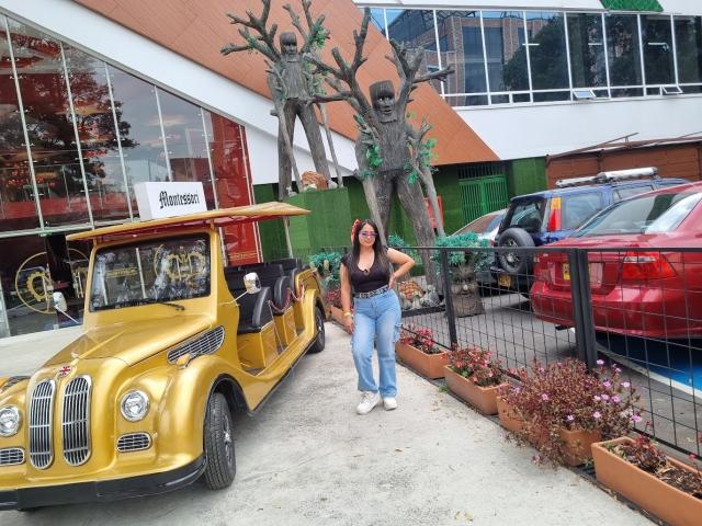 SofiiaThooner with big tits, wearing a black top and blue jeans, stands next to a golden jeep, her hand on her head, in a parking lot with a tree sculpture.