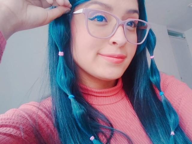 SofiiaThooner with blue braids, pink turtleneck, and purple glasses, touching her hair. She has blue eyes, pink lips, and a slight smile. Her tits are small.