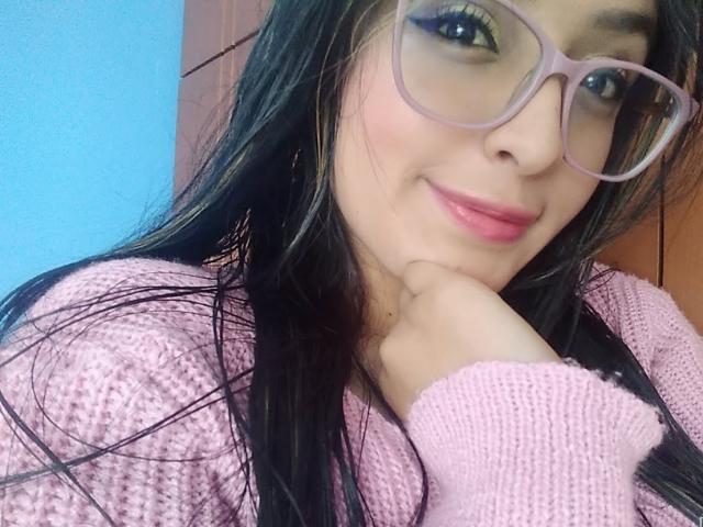 SofiiaThooner, avec ses longs cheveux noirs, ses lunettes roses, son rouge à lèvres rose et son pull rose en tricot, sourit doucement, se touche le menton, et a l'air à la fois mignonne et séduisante..