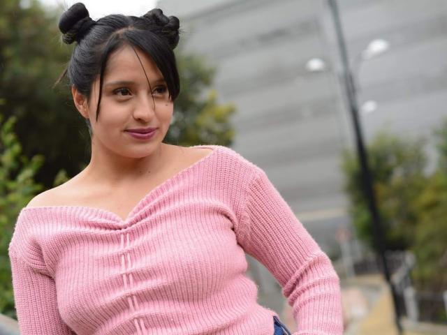 SofiiaThooner with black double buns, pink off-shoulder knit top, showing her tits, blue jeans, smiling slightly, standing outdoors, blurred background, overcast sky.