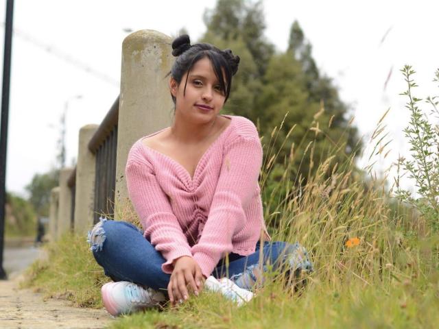 SofiiaThooner with black hair in double buns, pink cowl-neck sweater, ripped blue jeans, and pink-white sneakers, sitting cross-legged on grassy road beside concrete pillar.