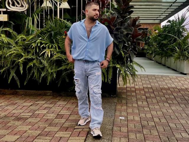 TonyLatino with a light blue shirt, ripped jeans, and white sandals, standing with hands in pockets, looking to the side, in a greenery-filled outdoor area.