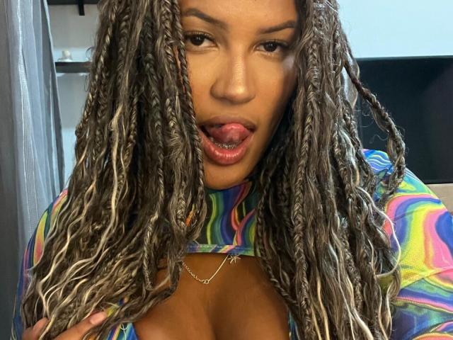 NoirSeduction69 with long, braided hair, tongue out, wearing a colorful, tie-front top, showing cleavage, wearing a delicate necklace, looking seductive, with a slight smirk.