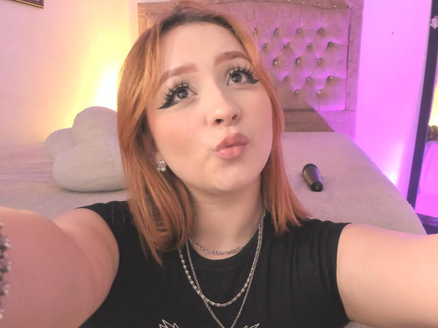 DarkCourtney with orange hair, black eyeliner, pouty lips, black shirt, necklace, and earrings, taking a selfie in a pink-lit bedroom, with a gray pillow and black dildo.